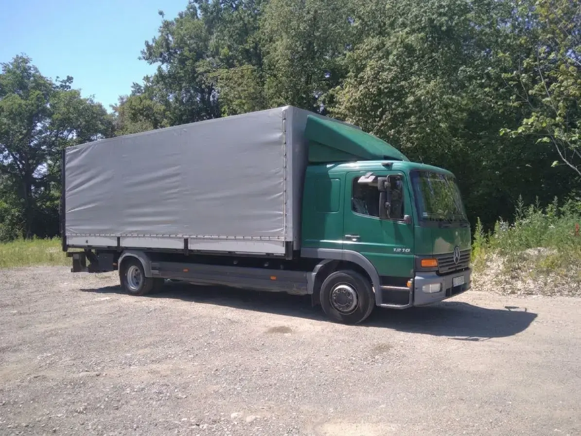 Zielona ciężarówka Mercedes-Benz Atego 1218 z szarą plandeką, widok z boku na żwirowym placu